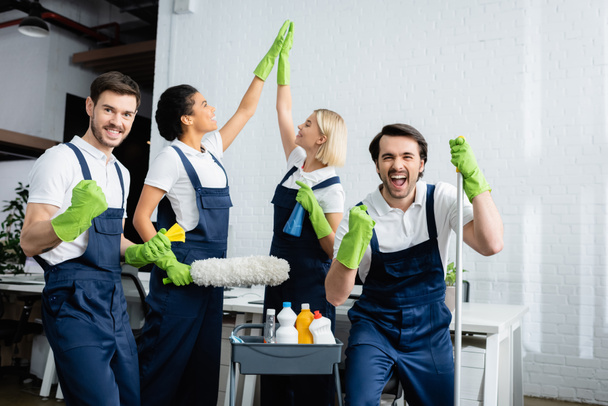 Emocionados limpiadores multiétnicos mostrando sí gesto y cinco altos en la oficina  - Foto, Imagen