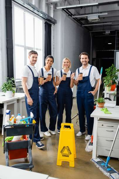 Cheerful multiethnic cleaners holding coffee to go near detergents in office  - Photo, Image