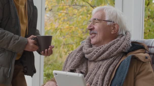 Verrouillage du vieil homme caucasien portant des vêtements chauds assis sur un fauteuil à bascule sur la terrasse et en utilisant une tablette. Puis son petit-fils adolescent lui apporte une tasse de thé chaud et ils commencent à parler - Séquence, vidéo
