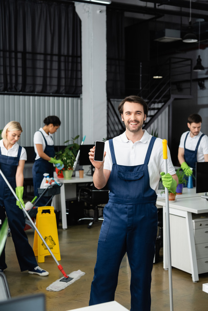 Χαμογελώντας εργαζόμενος της εταιρείας καθαρισμού κρατώντας smartphone με λευκή οθόνη στο γραφείο  - Φωτογραφία, εικόνα