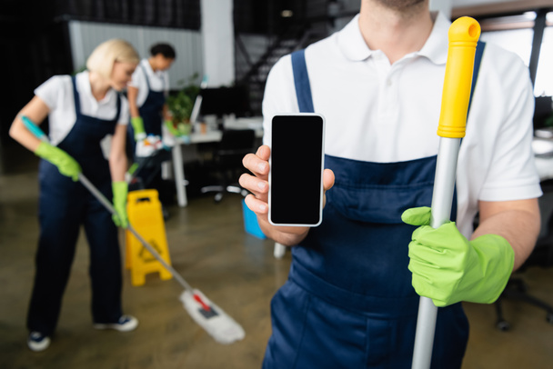 Limpiador sosteniendo teléfono inteligente y fregona en la oficina  - Foto, Imagen