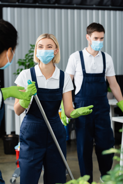 Interracial cleaners in medical masks and uniform standing in office  - Photo, Image