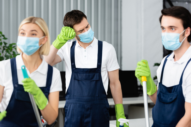 Trabajador cansado de servicio de limpieza en máscara médica de pie cerca de colegas en primer plano borroso en la oficina  - Foto, Imagen