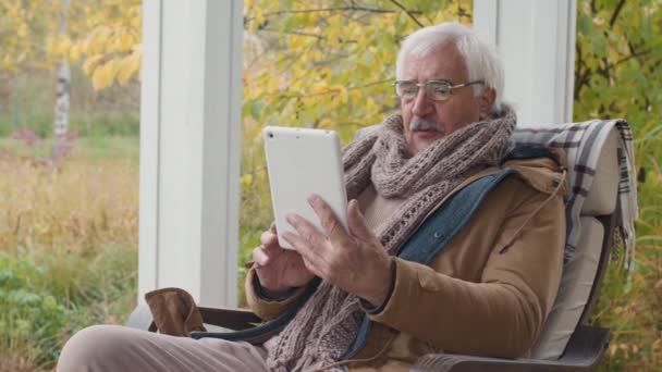 暖かい服と眼鏡を身に着けている高齢者の白人男性の中程度のショットテラスでロッキングチェアに座って、デジタルタブレットを使用して友人とビデオ会話をする - 映像、動画