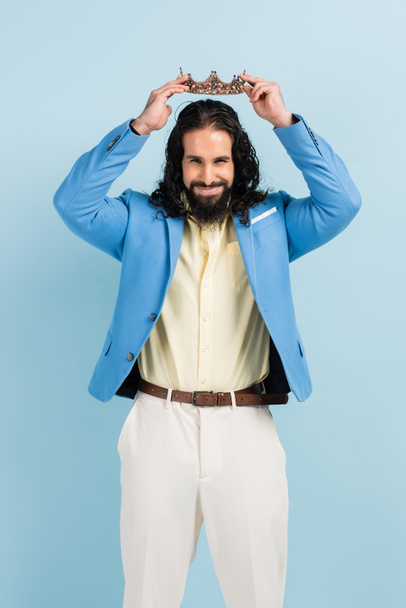 bearded hispanic man in jacket wearing crown and smiling isolated on blue - Photo, Image