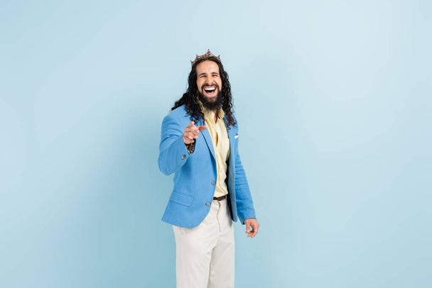 bärtiger hispanischer Mann in Krone und Jacke lacht, während er mit dem Finger auf blau zeigt - Foto, Bild