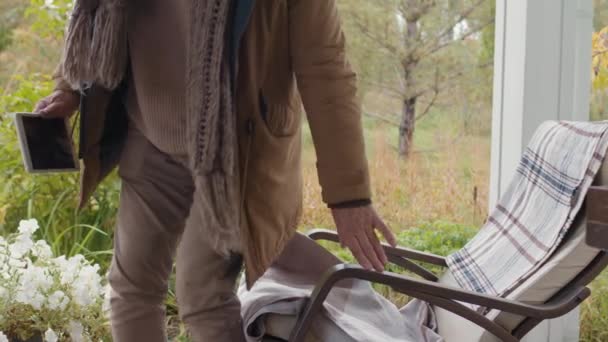 Aussperrung eines älteren kaukasischen Mannes mit warmer Kleidung und Brille, der sich auf einen Schaukelstuhl auf der Terrasse setzt und Videotelefonate per digitalem Tablet entgegennimmt - Filmmaterial, Video