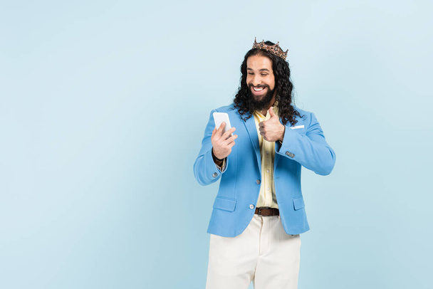 bearded hispanic man in jacket and crown showing thumb up during video call isolated on blue - Photo, Image
