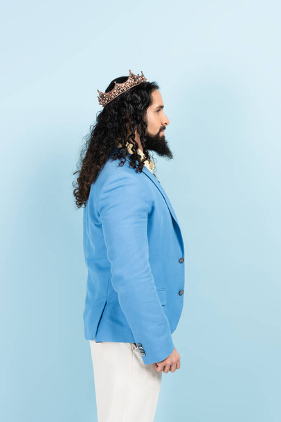 side view of bearded hispanic man in jacket and crown isolated on blue - Photo, Image
