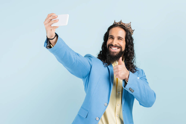 glücklicher hispanischer Mann in Jacke und Krone zeigt Daumen hoch und macht Selfie isoliert auf blauem Grund - Foto, Bild