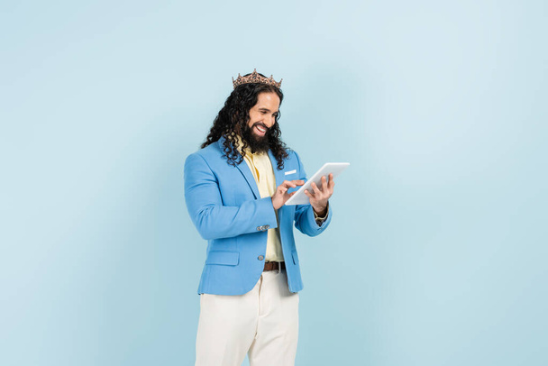 heureux hispanique homme en veste et couronne tenant tablette numérique isolé sur bleu - Photo, image