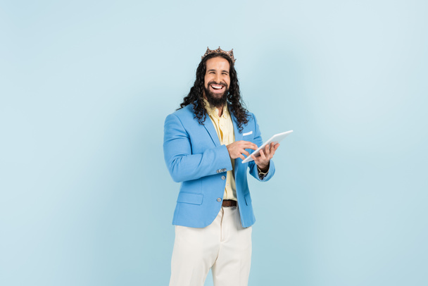 Χαρούμενος Ισπανόφωνος άνδρας με σακάκι και στέμμα κρατώντας ψηφιακό δισκίο απομονωμένο σε μπλε - Φωτογραφία, εικόνα
