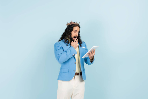 geschokte Spaanse man in jas en kroon met digitale tablet geïsoleerd op blauw - Foto, afbeelding