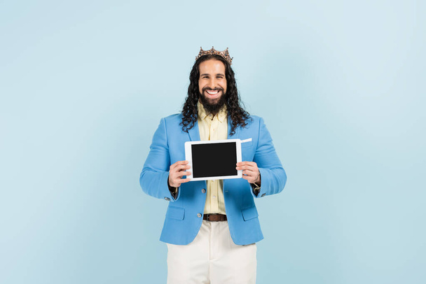 šťastný hispánec v bundě a koruně drží digitální tablet s prázdnou obrazovkou izolované na modré - Fotografie, Obrázek