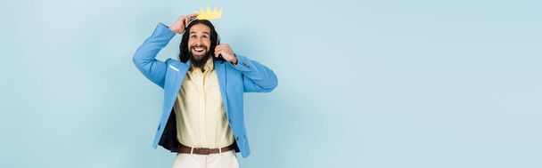 счастливый латиноамериканец в пиджаке держит бумажную корону на палочке изолированы на синий, баннер - Фото, изображение