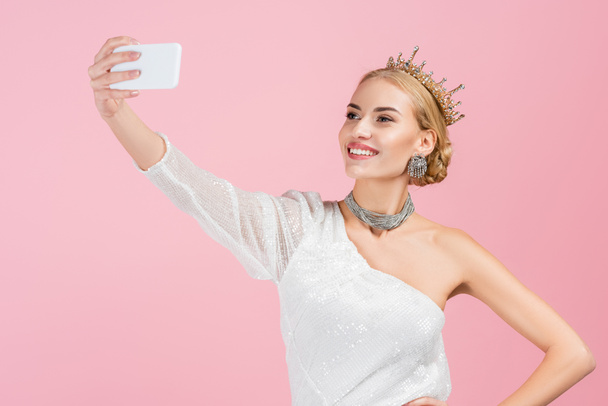 mulher loira em coroa de luxo tomando selfie no smartphone isolado em rosa - Foto, Imagem