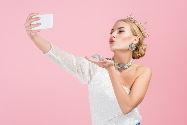 mulher loira em coroa de luxo tomando selfie no smartphone e enviando beijo de ar isolado no rosa - Foto, Imagem