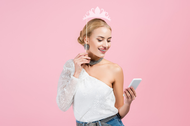 счастливая женщина держит бумажную корону на палочке и смартфон изолирован на розовый - Фото, изображение