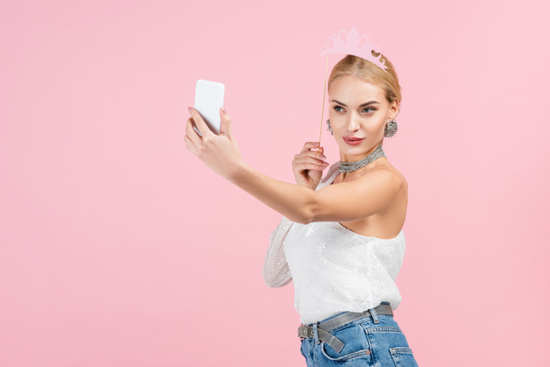 szőke nő gazdaság papír korona boton, és selfie okostelefon elszigetelt rózsaszín - Fotó, kép