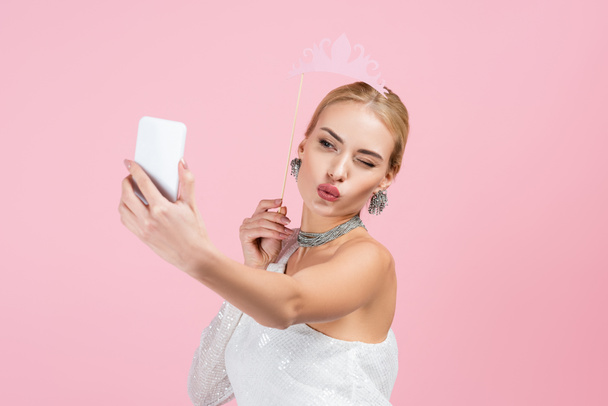 blond vrouw met papier kroon op stok en het nemen van selfie terwijl het verzenden van lucht kus geïsoleerd op roze - Foto, afbeelding