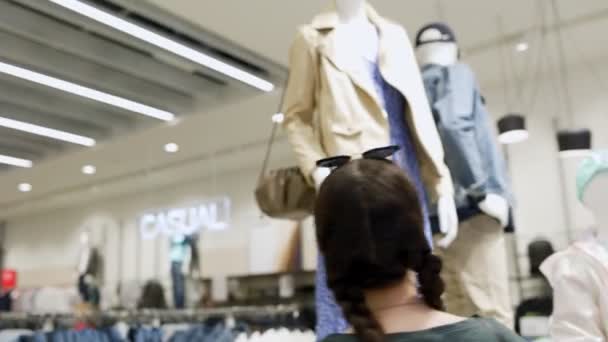 Una giovane donna caucasica guarda un manichino con vestiti in un negozio. In tempo reale. Il concetto di consumismo e shopping. - Filmati, video