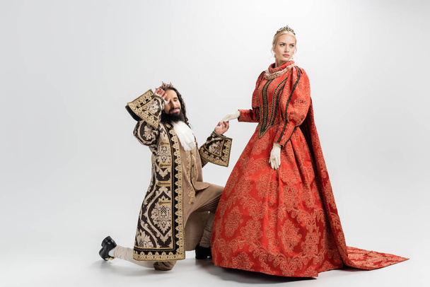 rey hispano en corona y ropa medieval de pie sobre la rodilla mientras toma de la mano de la reina rubia en blanco - Foto, Imagen