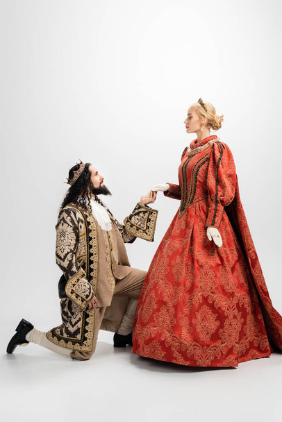 hispanic king in crown and medieval clothing standing on knee and holding hand of blonde queen on white - Photo, Image