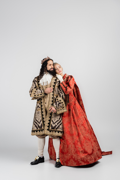 longitud completa de la reina rubia en corona real apoyada en el rey hispano en ropa medieval en blanco - Foto, Imagen