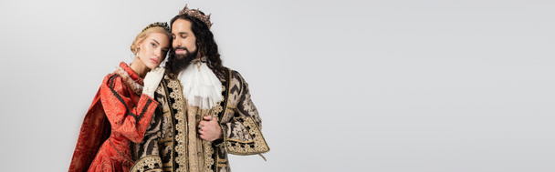 reina rubia en corona real apoyada en rey hispano en ropa medieval aislada en gris, bandera - Foto, Imagen