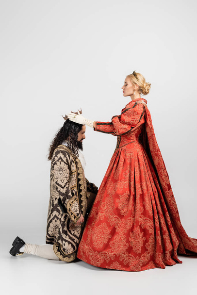 πλαϊνή άποψη της ξανθιάς βασίλισσας φορώντας στέμμα στο ισπανικό βασιλιά με μεσαιωνικά ρούχα στέκεται στα γόνατα σε λευκό - Φωτογραφία, εικόνα