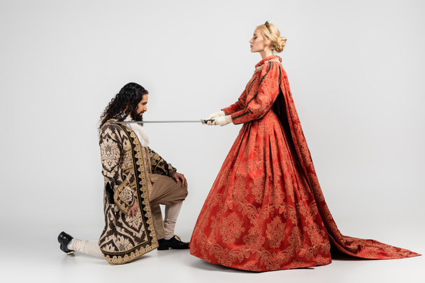 widok z boku blondynka królowa trzyma miecz w pobliżu hiszpańskiego króla w średniowiecznej odzieży stojącej na kolanie na białym - Zdjęcie, obraz