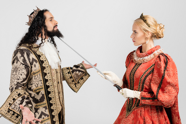 blonde queen holding sword near neck of hispanic king in medieval clothing and crown isolated on white - Photo, Image