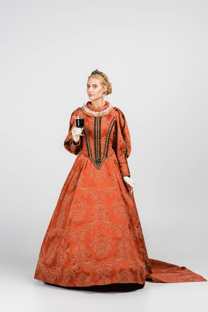 full length of queen in dress and crown holding glass of red wine on white  - Photo, Image