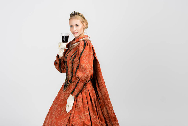 βασίλισσα με φόρεμα και χρυσό στέμμα κρατώντας γυαλί και πίνοντας κόκκινο κρασί σε λευκό  - Φωτογραφία, εικόνα