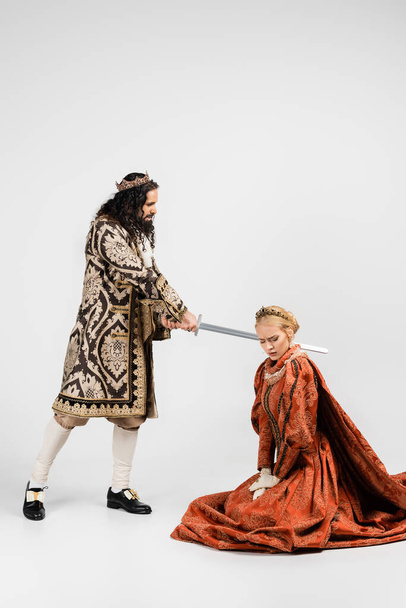 Orta çağ giysili zalim İspanyol kralın elinde beyaz taç giymiş korkmuş sarışın kraliçenin yanında. - Fotoğraf, Görsel