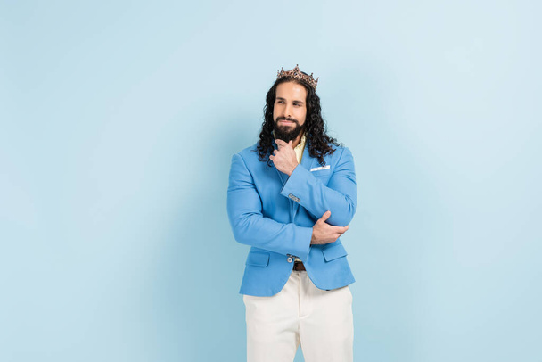 nachdenklicher und bärtiger hispanischer Mann in Krone und Jacke posiert auf blauem Grund - Foto, Bild