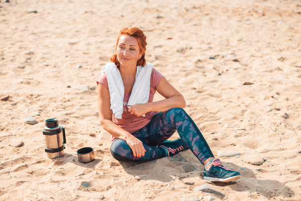 Dopo l'allenamento. Donna adulta sorridente in abiti sportivi, seduta sulla sabbia. Accanto ad essa c'è una bottiglia di nutrizione sportiva. Il concetto di stile di vita sportivo e nutrizione sportiva. - Foto, immagini
