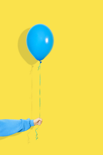 女性の手は青い気球を保持します。党や現在の概念。青い風船と黄色の背景に隔離された青いパーカーの手. - 写真・画像