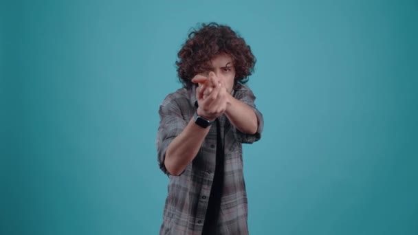Charismatic hipster imitates a pistol with his hands, and shoots in front, dressed in a plaid shirt, with curly hair, in the studio on a turquoise background. - Footage, Video