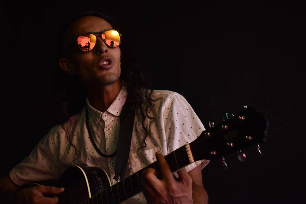 Bello uomo capelli ricci lunghi che suona la chitarra in modo bohemien con occhiali da sole, stile rockstar - Foto, immagini