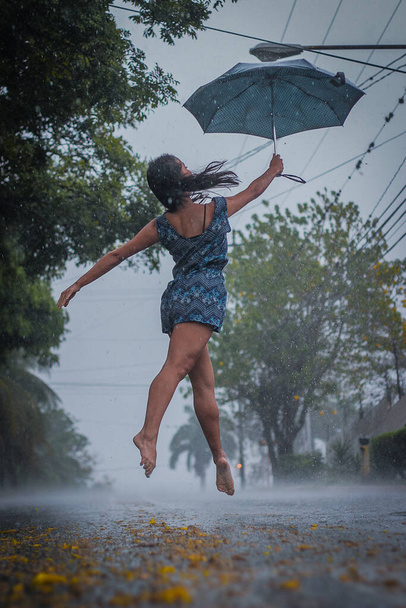 Kobieta artysta tancerka w ubraniu casual wykonuje na ulicy szczęśliwie i swobodnie w deszczu. Skocz boso po ulicy z szarym parasolem - Zdjęcie, obraz