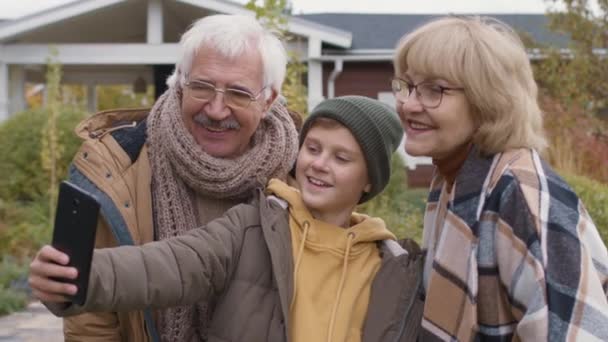 Średnie ujęcie starszej białej kobiety, jej męża i nastoletniego wnuka w ciepłych, luźnych ubraniach stojących przed domem na wsi i robiących selfie za pomocą aparatu telefonicznego - Materiał filmowy, wideo