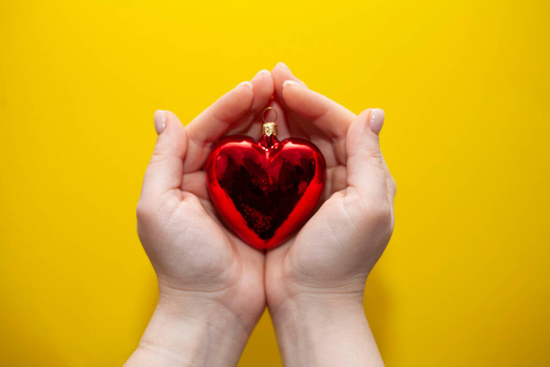 Le mani femminili su uno sfondo giallo tengono un giocattolo di albero di Natale nella forma di un cuore - Foto, immagini