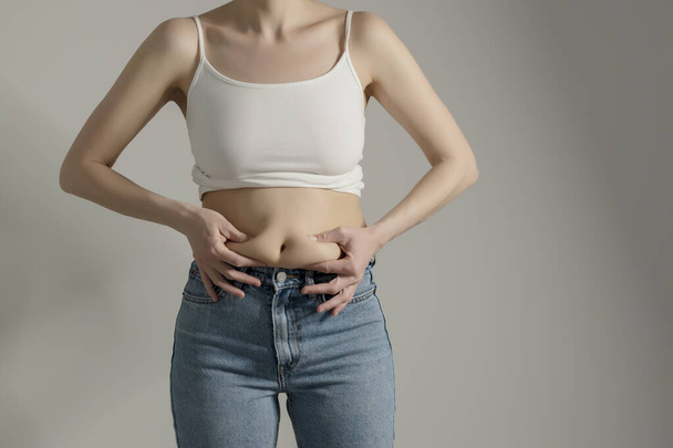 Woman in jeans and white shirt squeezing her belly fat. Woman`s hips closeup raw studio shot in grey background. Dieting and fat loss concept. - Photo, image