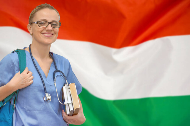 ハンガリーの国旗を背景に聴診器と本を手にした女子学生の医師。医学教育の概念。ハンガリーで医学を学ぶ - 写真・画像