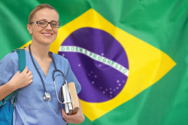 ブラジルの旗の背景に手に聴診器と本を持つ女性学生の医師。医学教育の概念。ブラジルで医学を学ぶ - 写真・画像