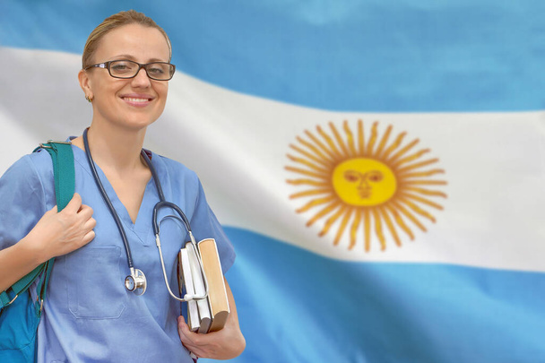 アルゼンチンの旗の背景に手に聴診器と本を持つ女性学生の医師。医学教育の概念。アルゼンチンで医学を学ぶ - 写真・画像