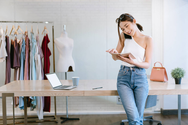 Beautiful Female Entrepreneur. Young cheerful Asian Woman fashion designer talking on mobile phone and checking notes in notebook. Startup Small business owner, Freelance work., SME marketing concept - Zdjęcie, obraz