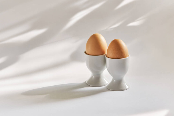 Zwei braune Eier in Eierbechern auf weißem Hintergrund mit einem schönen Schatten - Foto, Bild