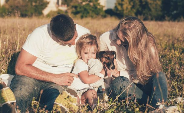 семья с маленькой дочкой сидят на траве в парке и играют с собакой - Фото, изображение